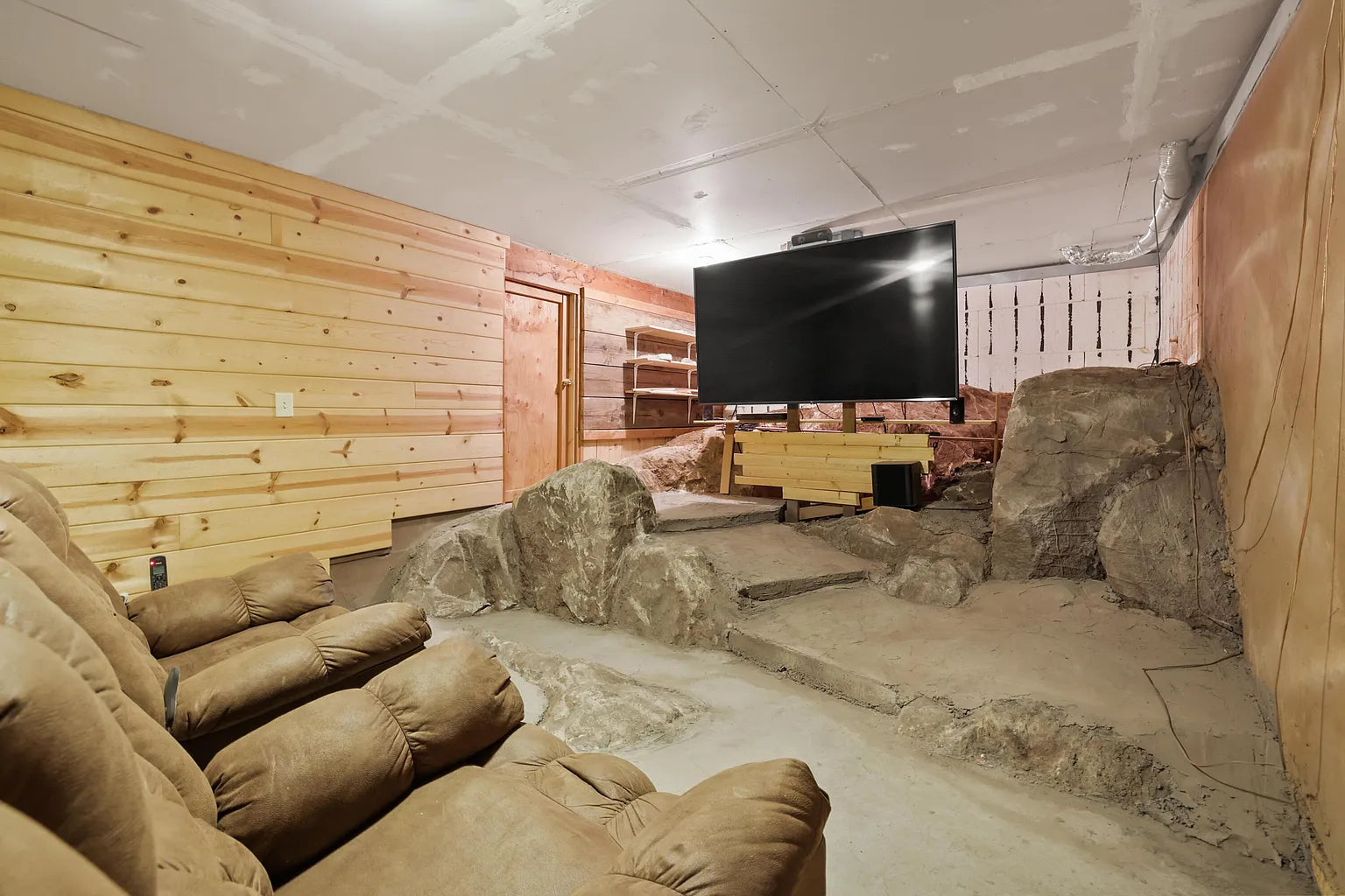 A home theater room with recliner chairs, wood-paneled walls, and a large TV on a stand. The floor features natural rock formations, giving the room a cave-like appearance.