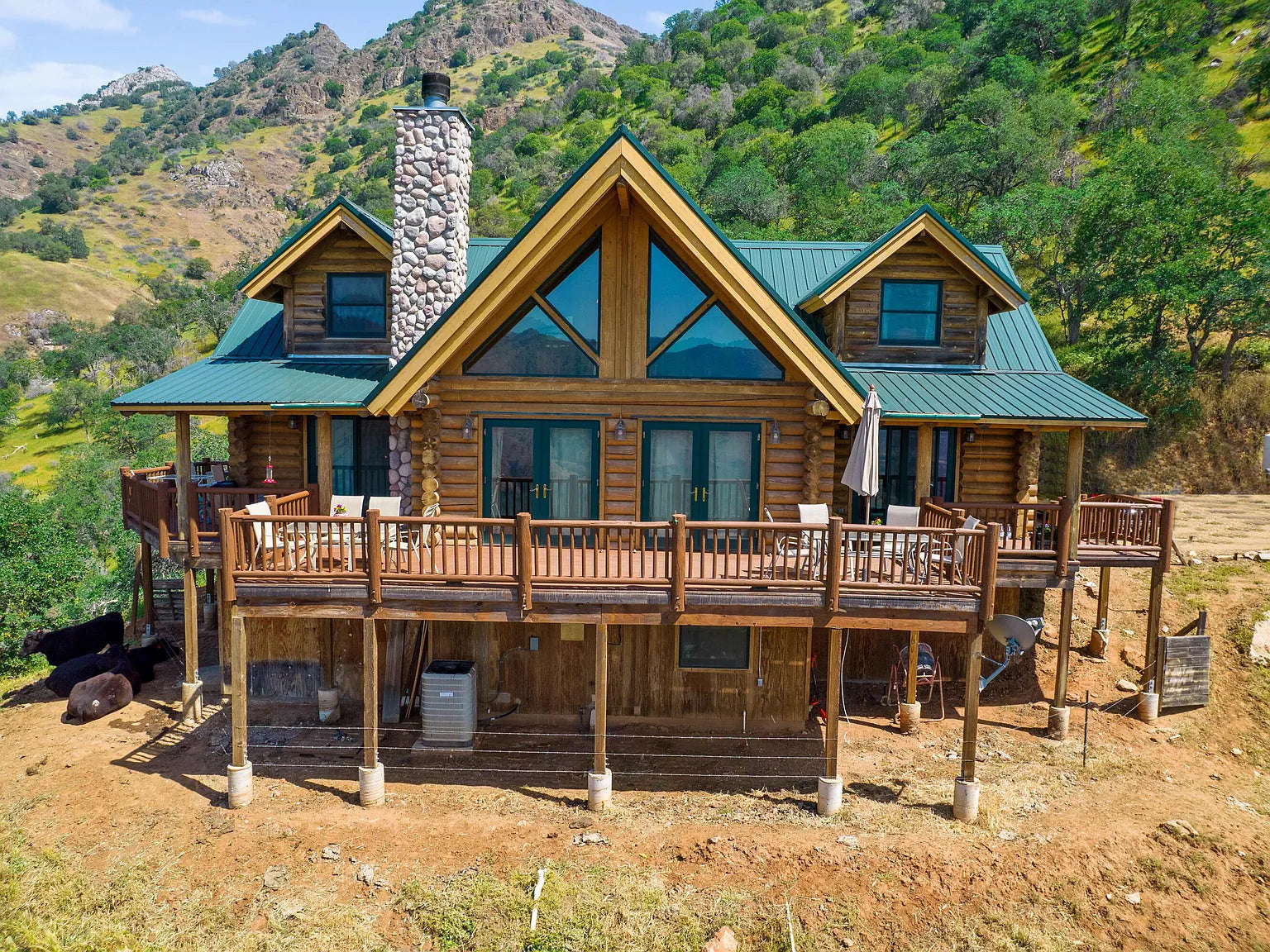 A large log cabin with a green metal roof, stone chimney, and wide wooden deck sits on a hillside surrounded by green trees and mountains under a blue sky.