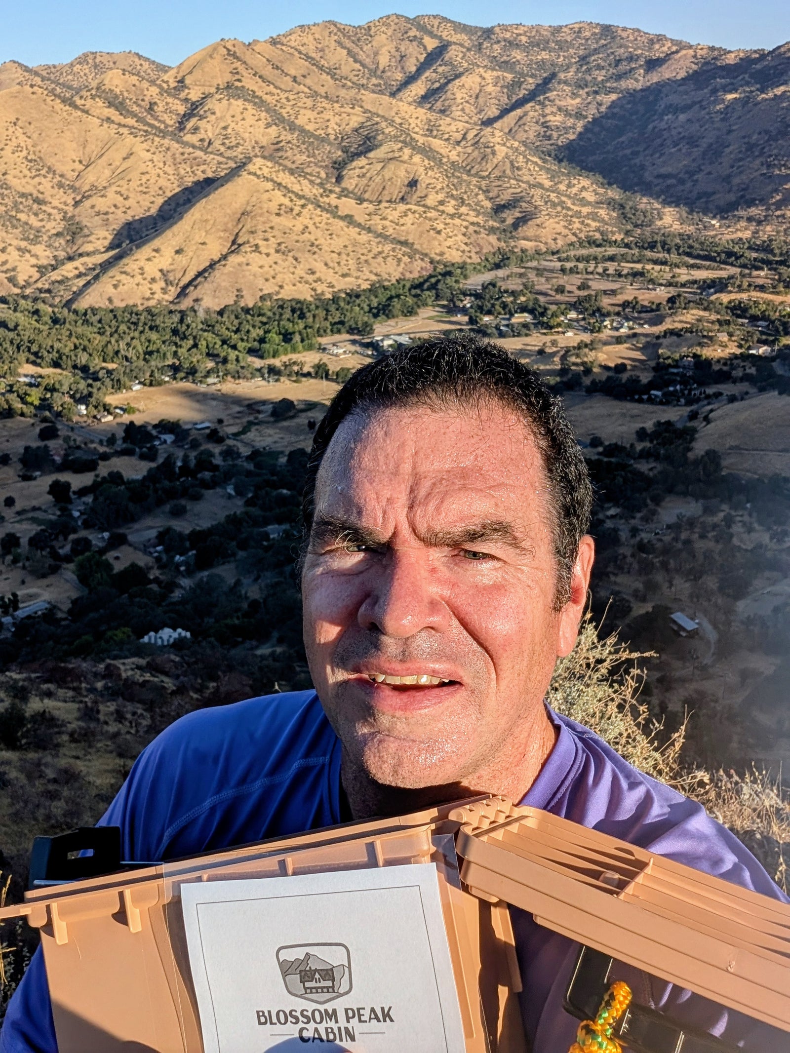 A man stands on a hillside with a scenic view of a valley and mountains behind him, holding a sign that reads Blossom Peak Cabin. The sun is shining, casting clear shadows and illuminating the landscape.