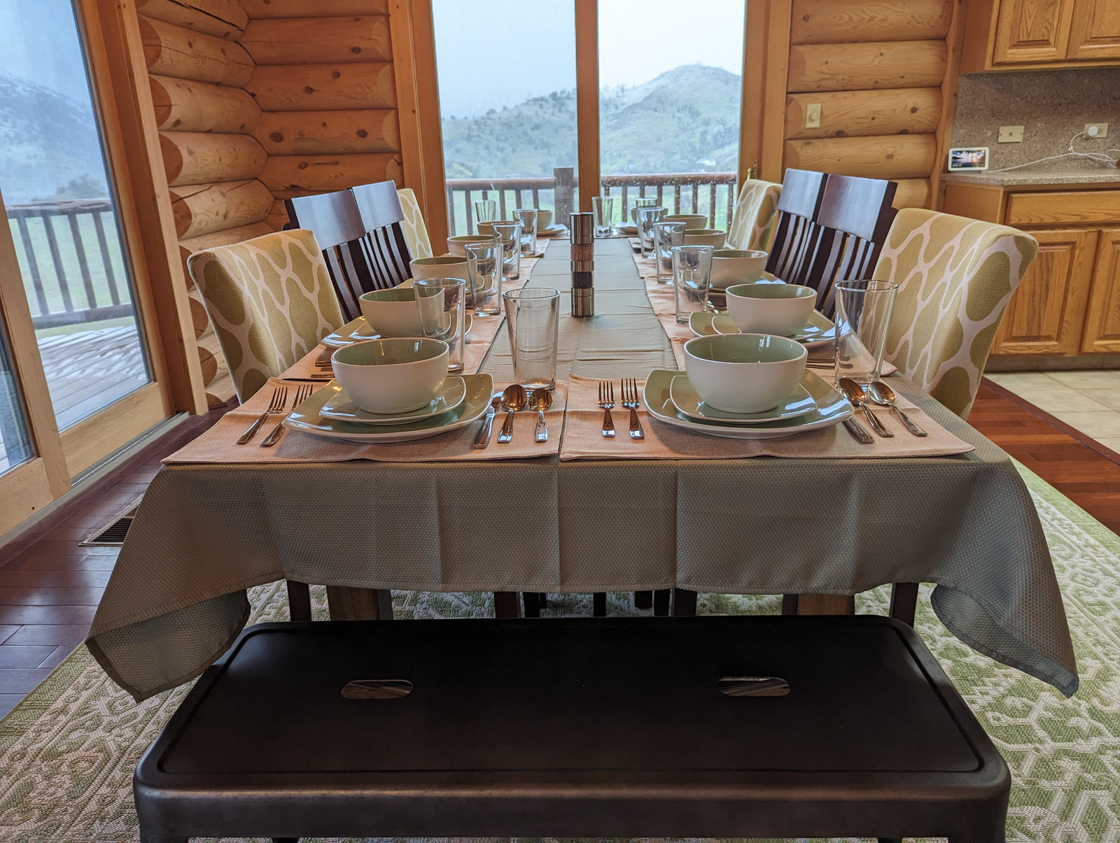 A dining table set for eight with plates, bowls, cups, glasses, and silverware, surrounded by patterned chairs and a bench, in a log cabin with large windows overlooking mountains.