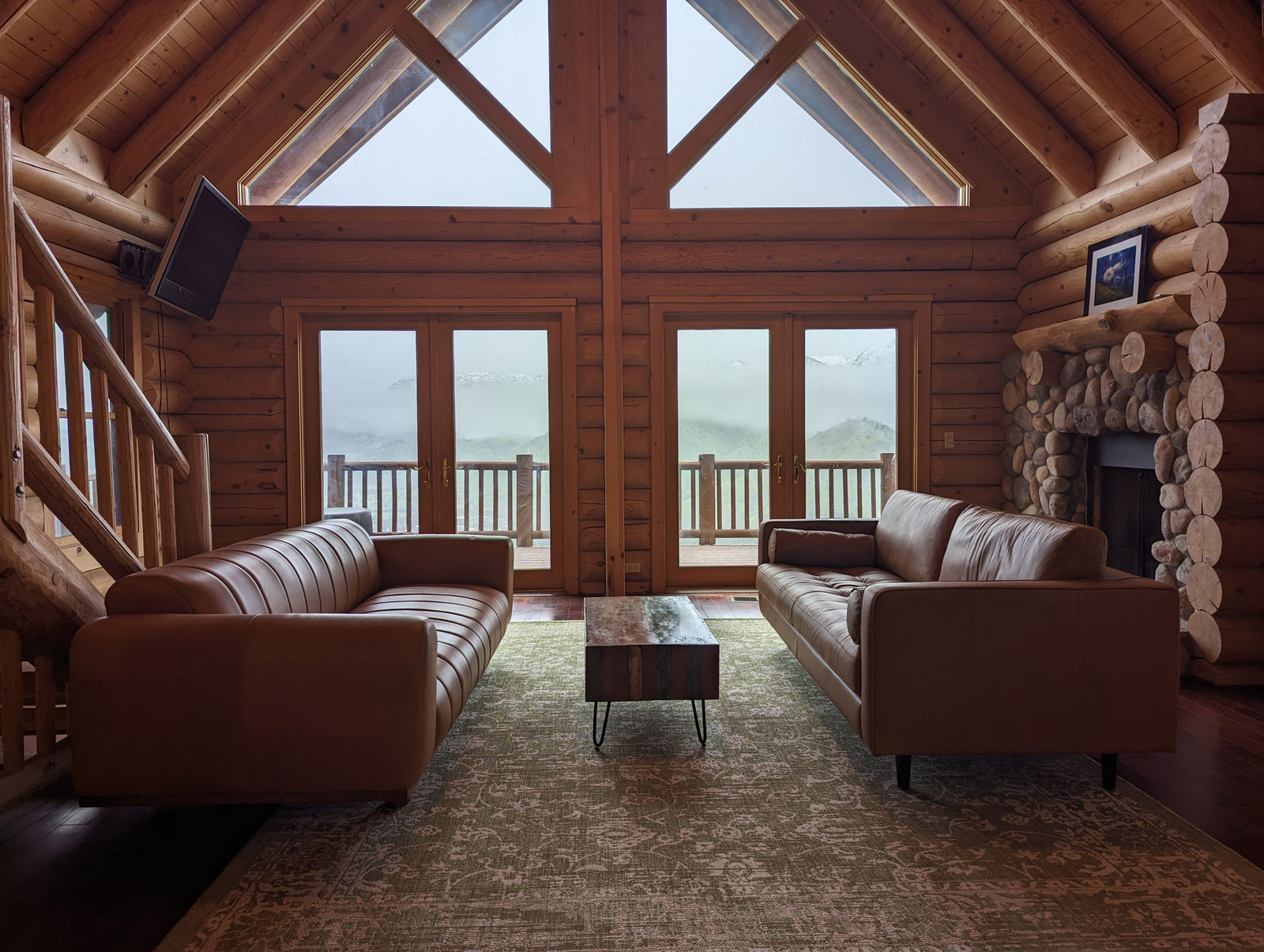 A cozy log cabin living room with two brown leather sofas facing each other, a wooden coffee table between them, large windows with a mountain view, and a stone fireplace on the right.