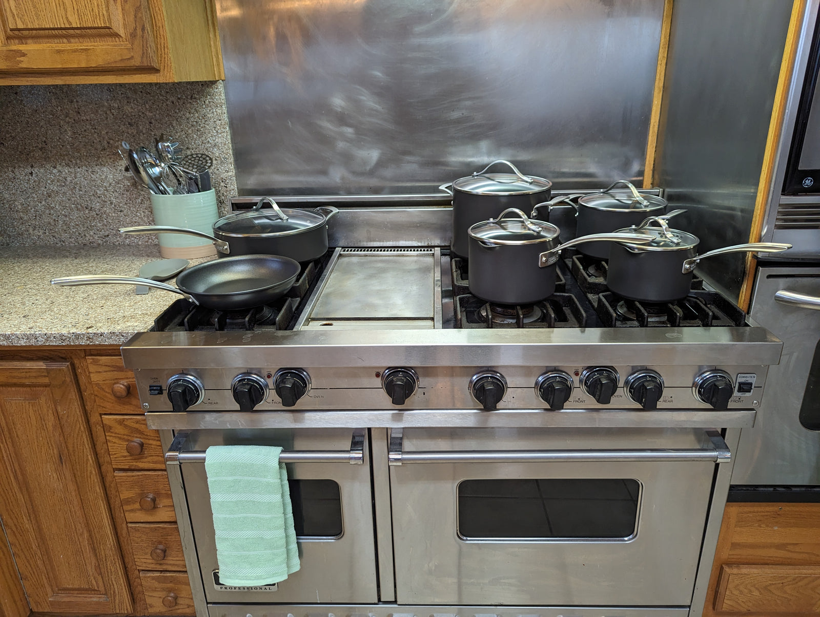 A stainless steel stove with six black pots and pans on the burners. The stove has two ovens below and a green-striped towel hanging on the front handle. Wooden cabinets and kitchen utensils are in the background.