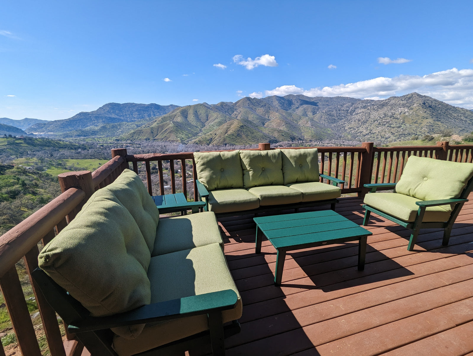 Outdoor wooden deck with green cushioned sofas and chairs, overlooking scenic mountains and a valley under a clear blue sky with a few clouds.