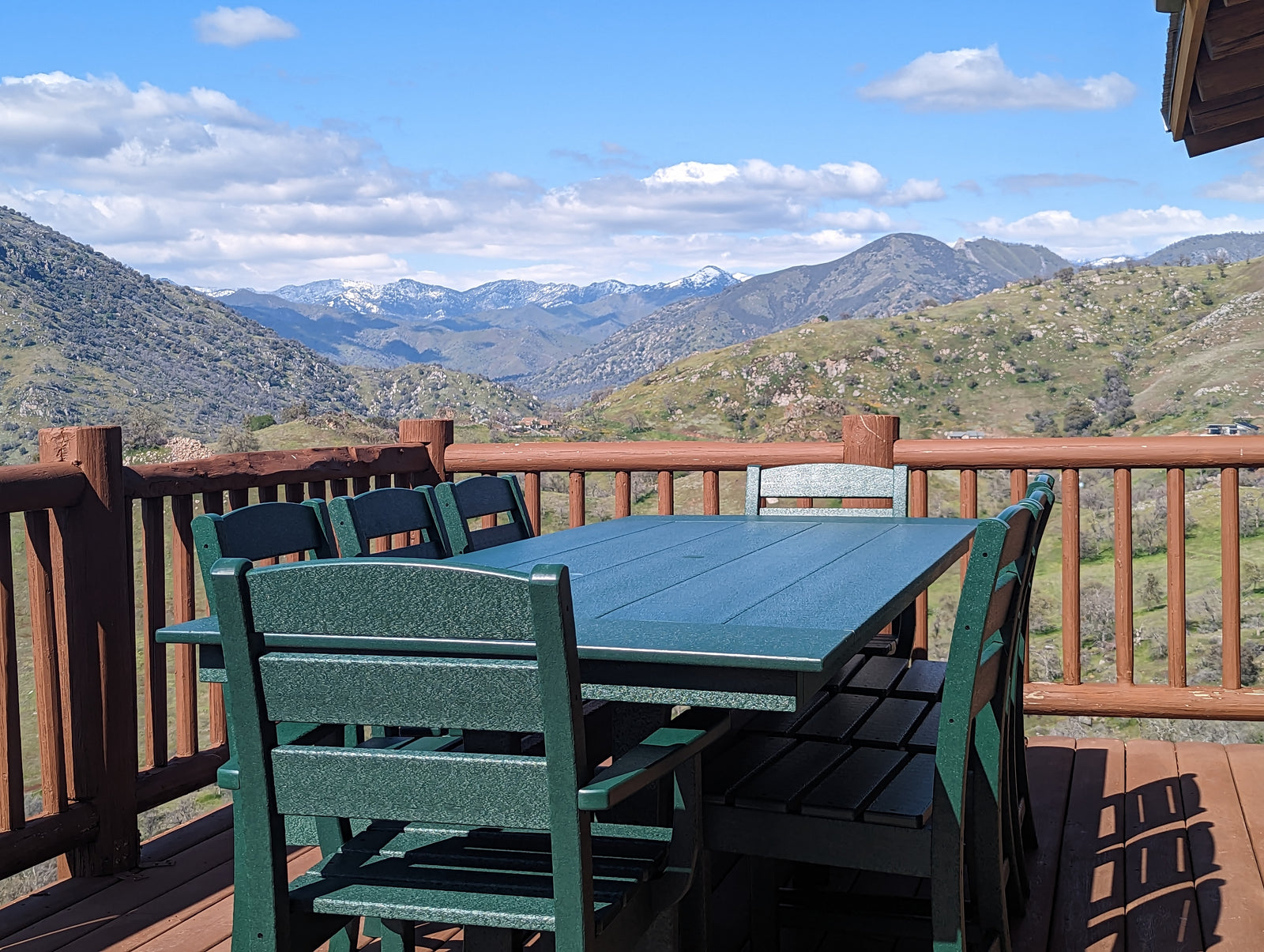 A green outdoor table with matching chairs sits on a wooden deck, overlooking rolling green hills and snow-capped mountains under a partly cloudy sky.