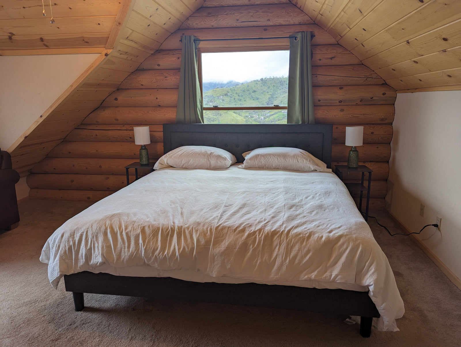 A cozy loft bedroom with a king-size bed, white bedding, and two pillows sits against a wooden log wall. Two side tables with lamps flank the bed, and a window with green curtains shows a scenic outdoor view.